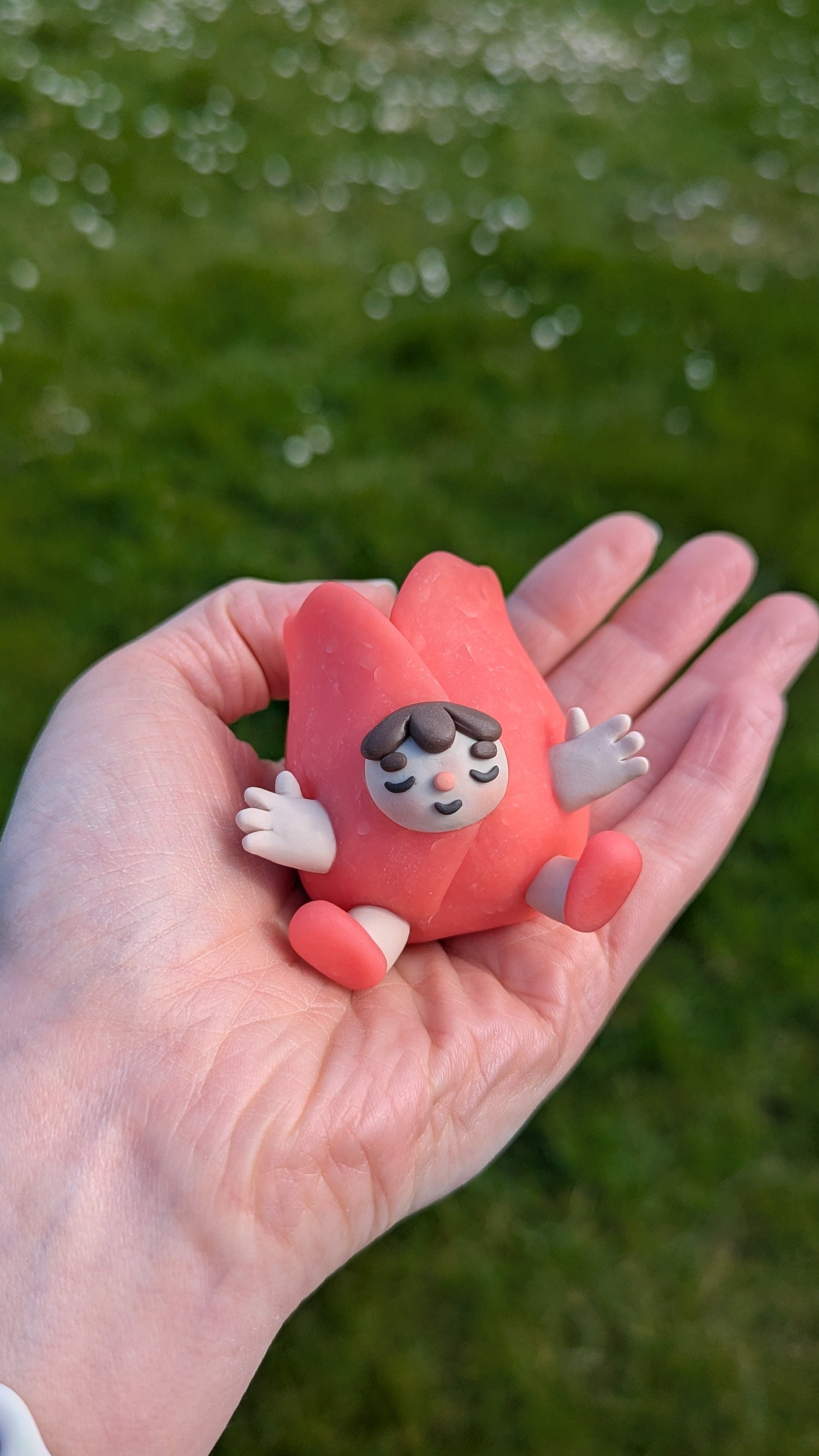 Hand holding a small red figurine with a face against a grassy background